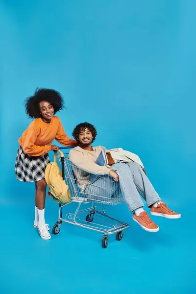 Two young women of different ethnicities are having fun sitting in a shopping cart against a blue backdrop in a studio setting. — Stock Photo