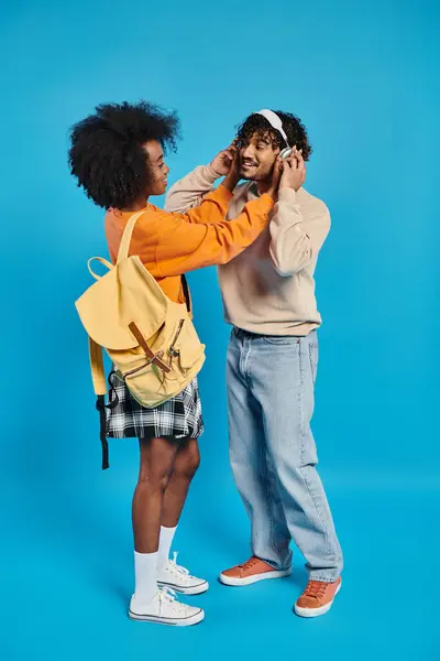 Un hombre y una mujer, ambos estudiantes interracial, de pie juntos en atuendo casual, con la mujer usando una mochila, en un contexto azul. - foto de stock