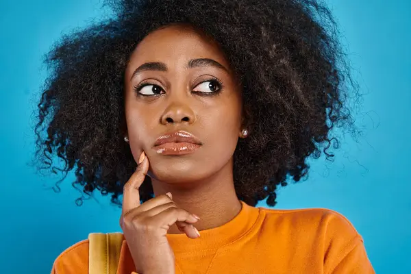 Ein stylisches afroamerikanisches College-Mädchen blickt mit ihrem atemberaubenden Afro-Haar vor blauem Hintergrund in einem Atelier nach oben. — Stockfoto