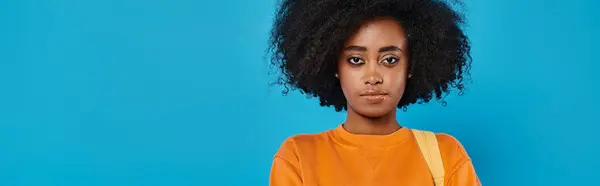 Una donna afro-americana elegante in abbigliamento casual si erge con fiducia con i suoi voluminosi capelli afro, di fronte a un vibrante sfondo blu. — Foto stock