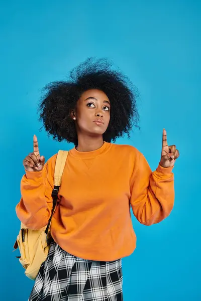 Chica universitaria afroamericana con peinado afro apuntando hacia arriba n telón de fondo azul en el estudio. - foto de stock
