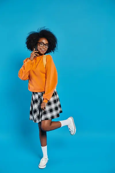 A stylish African American college girl stands confidently in a bright orange sweater and trendy plaid skirt against a blue backdrop. — Stock Photo