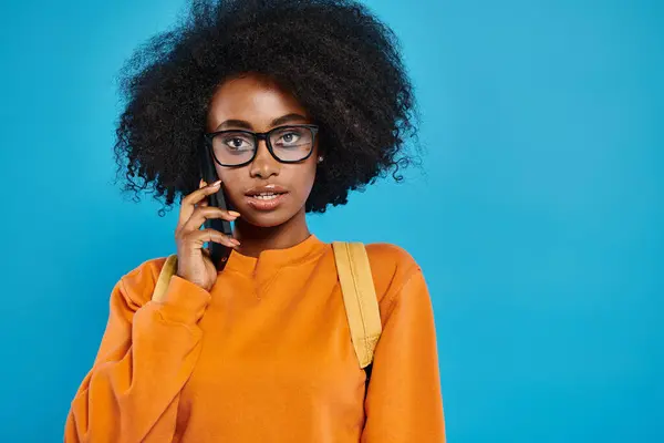 Una chica universitaria afroamericana con gafas chats en un teléfono celular contra un telón de fondo azul en un entorno de estudio. - foto de stock