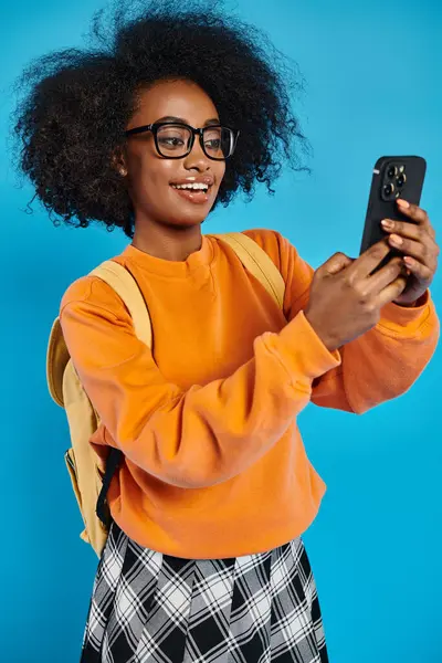 Une jeune afro-américaine en tenue décontractée prend un selfie avec son téléphone portable tout en portant un sac à dos, sur fond bleu dans un studio. — Photo de stock