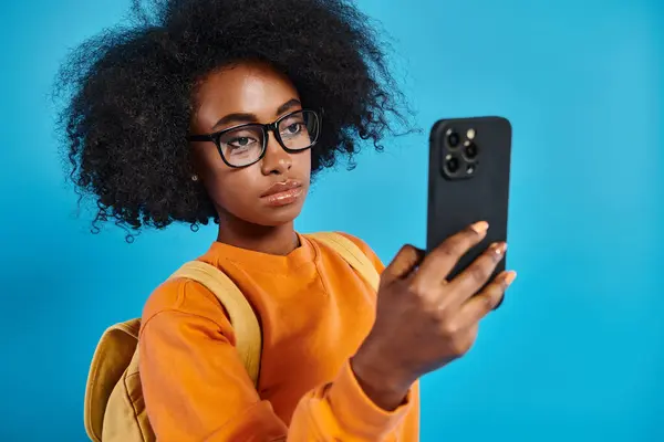 Una chica universitaria afroamericana con atuendo casual, usando gafas, tomando una selfie con su teléfono celular en un telón de fondo azul en un estudio. - foto de stock