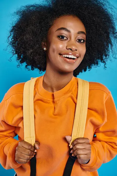A joyful African American college girl with an afro hairstyle smiles while holding a pair of suspenders, standing in casual attire against a blue backdrop in a studio. — Stock Photo