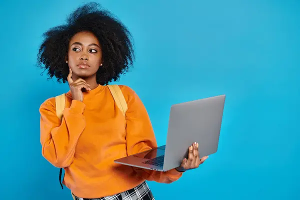African American college girl in casual attire standing with laptop in front of blue background. — Stock Photo