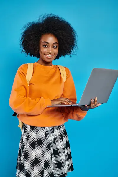 Una chica universitaria afroamericana sonríe mientras sostiene una computadora portátil en un estudio con un telón de fondo azul. - foto de stock