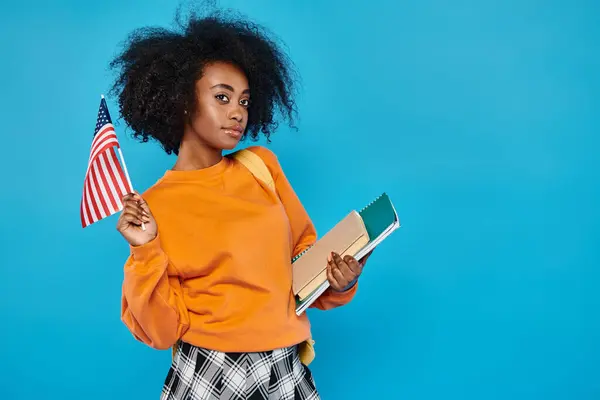 Una joven universitaria afroamericana de pie con orgullo, sosteniendo un libro y una bandera americana. - foto de stock