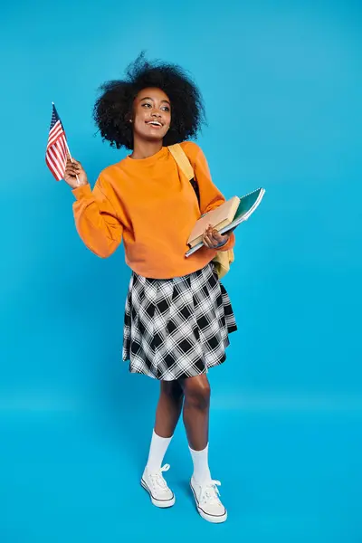 Una universitaria afroamericana de pie con un libro en una mano y una bandera americana en la otra, exudando patriotismo. - foto de stock