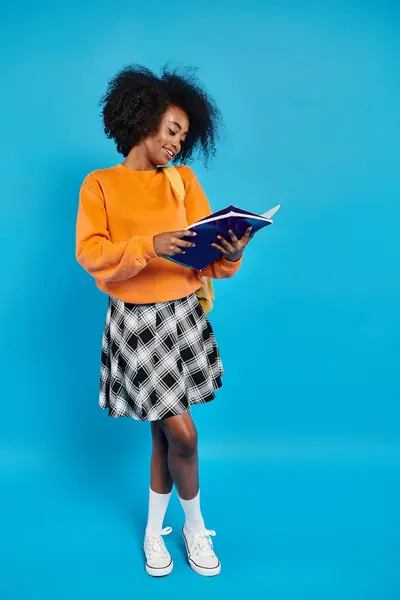 Uma menina universitária afro-americana, de traje casual, está contra um pano de fundo azul, envolta em um livro. — Fotografia de Stock
