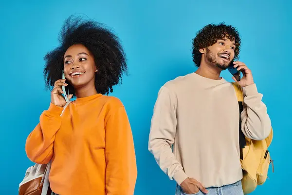 A man and a woman, interracial students, standing together in casual attire, engaged in conversation on cell phones. Blue backdrop in studio. — Stock Photo