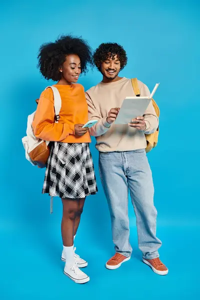 Una pareja interracial de estudiantes parados uno al lado del otro en atuendo casual sobre un fondo azul en un estudio. - foto de stock