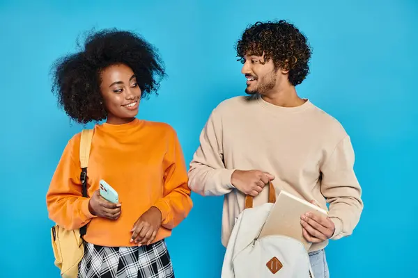 Un hombre y una mujer, estudiantes interraciales, se unen en atuendo casual contra un telón de fondo azul en un estudio. - foto de stock