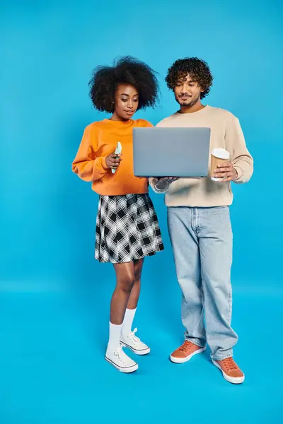 An interracial couple, standing together, holding a laptop against a blue backdrop. — Stock Photo