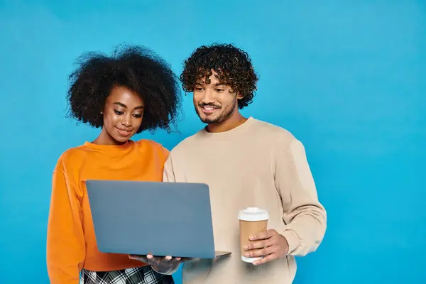 Un homme et une femme, tenant un ordinateur portable et une tasse de café, debout ensemble dans un studio avec une toile de fond bleue. — Photo de stock