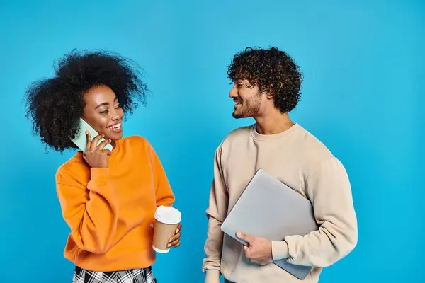 An interracial couple of students standing together in casual attire against a blue backdrop, using devices — Stock Photo