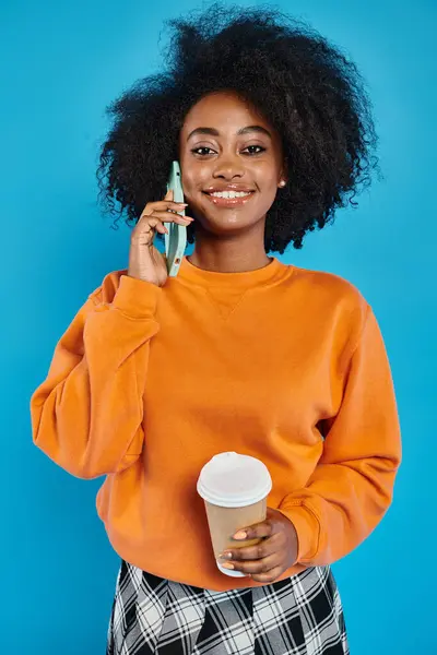 Una mujer con estilo de diferentes etnias sosteniendo una taza de café mientras habla en un teléfono celular contra un telón de fondo azul. - foto de stock