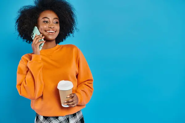 Una donna di colore che tiene una tazza di caffè mentre è impegnata in una conversazione telefonica, sullo sfondo blu. — Foto stock