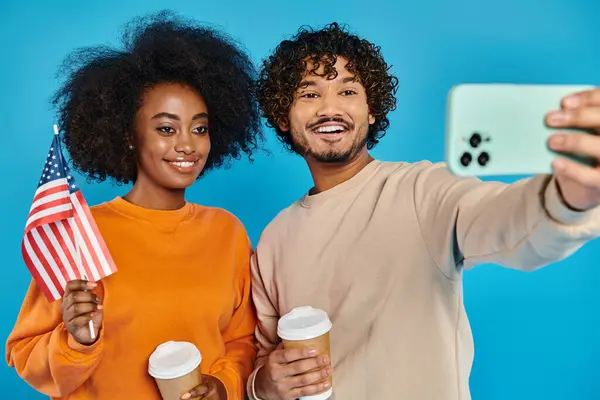 An interracial man and woman in casual attire take a selfie with American flag against a blue backdrop in a studio. — Stock Photo