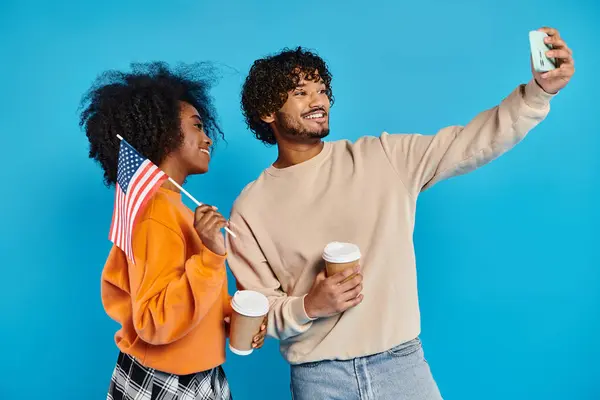 Um casal inter-racial, elegantemente vestido, capturando um momento junto com uma selfie de telefone celular contra um pano de fundo azul, bandeira americana — Fotografia de Stock