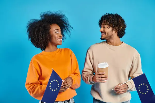 Um casal inter-racial de estudantes que estão lado a lado em trajes casuais com bandeiras da UE contra um pano de fundo azul em um estúdio. — Fotografia de Stock