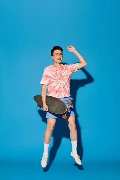 Un jeune homme élégant et beau en tenue tendance prend une pose, tenant un skateboard dans sa main droite sur un fond bleu. — Photo de stock