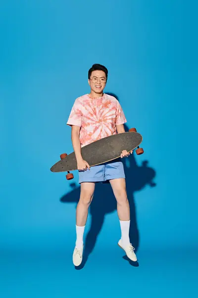 Uomo alla moda in abbigliamento alla moda che tiene lo skateboard in mano su sfondo blu. — Foto stock