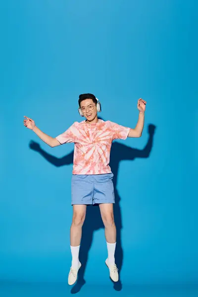 Un jeune homme à la mode en tenue tendance saute joyeusement dans les airs avec les mains levées sur un fond bleu. — Photo de stock