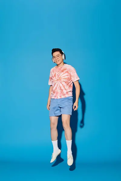 A stylish young man poses actively in trendy attire, wearing headphones against a blue backdrop. — Stock Photo