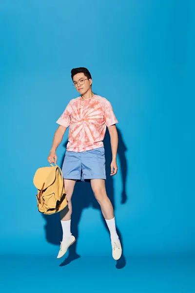 A stylish young man in a tie-dyed shirt energetically holds a backpack against a vibrant blue backdrop. — Stock Photo