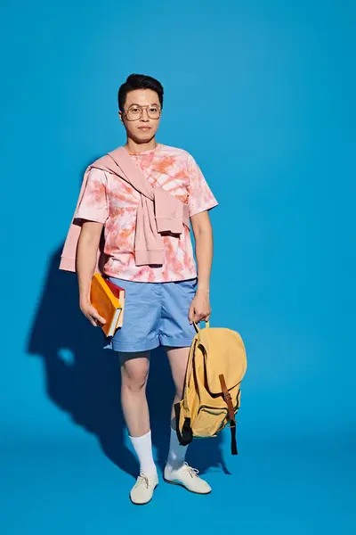 Stylish young man in pink shirt and blue shorts energetically holding a yellow bag against a blue backdrop. — Stock Photo