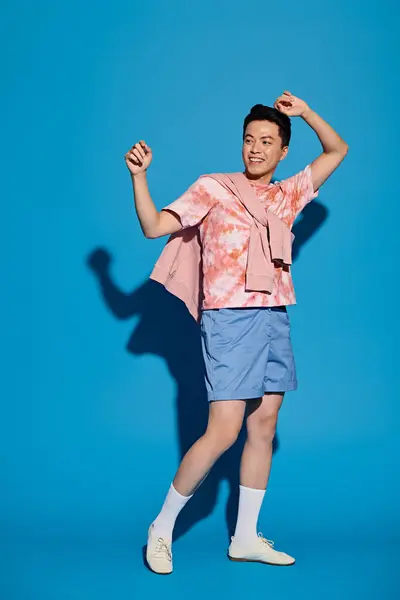 Un jeune homme élégant en chemise rose et short bleu pose énergiquement sur fond bleu, mettant en valeur des vêtements à la mode. — Photo de stock