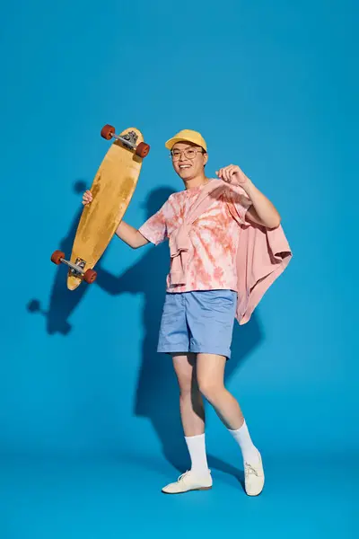 Um jovem elegante em trajes de moda posa energeticamente, segurando um skate, contra um cenário azul vibrante. — Fotografia de Stock