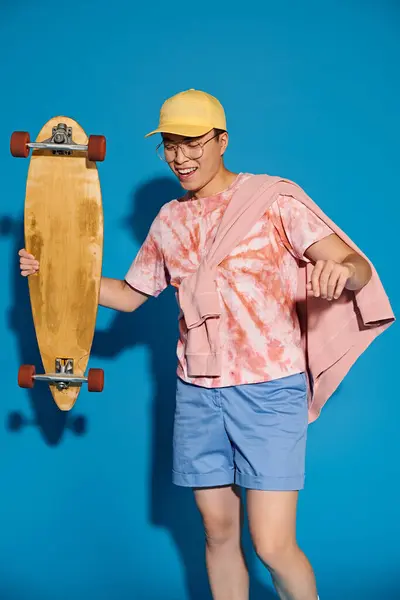 Young man in trendy attire confidently holds skateboard in front of vibrant blue wall. — Stock Photo
