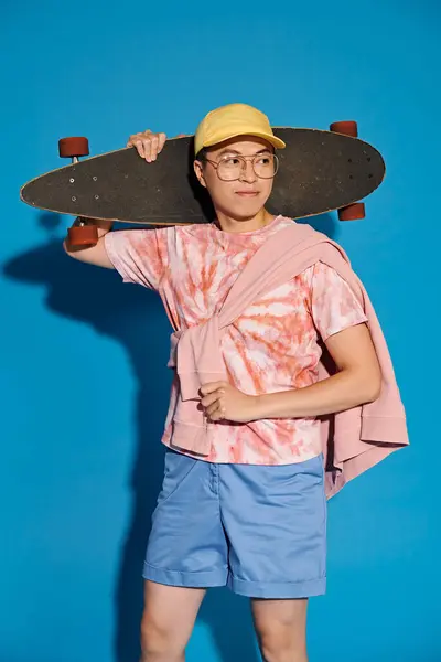 A stylish young man effortlessly holds a skateboard on his head against a blue backdrop. — Stock Photo