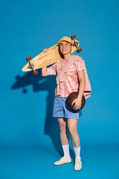 A stylish young man in trendy attire effortlessly balances a skateboard and a ball against a vibrant blue backdrop. — Stock Photo