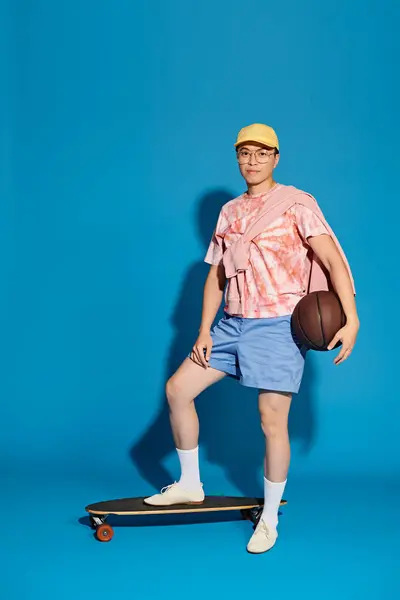 Un joven con estilo se equilibra en un monopatín mientras sostiene una pelota de baloncesto, mostrando su habilidad y confianza contra un telón de fondo azul. - foto de stock