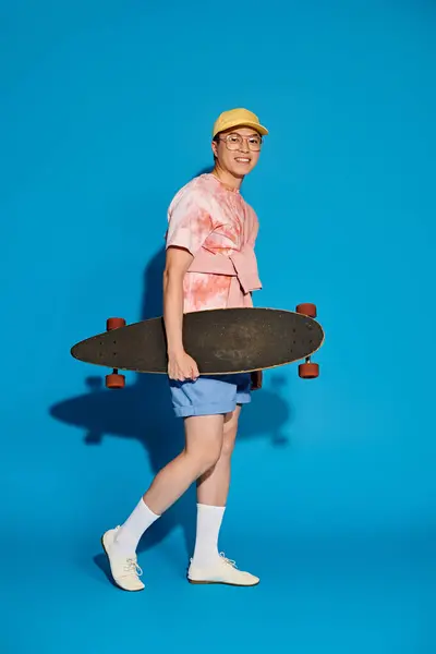 Un giovane elegante in abiti alla moda tiene uno skateboard su uno sfondo blu vibrante. — Foto stock