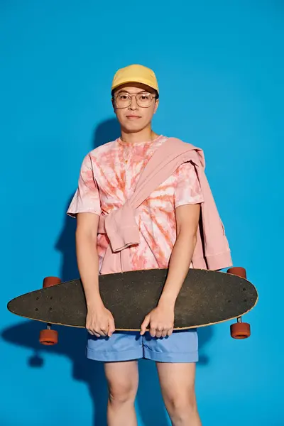 Un giovane uomo alla moda in abbigliamento elegante posa con fiducia con uno skateboard su uno sfondo blu vibrante. — Foto stock