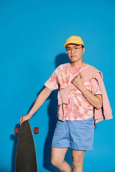 A trendy young man poses with a skateboard in front of a vibrant blue wall, showcasing urban style and energy. — Stock Photo