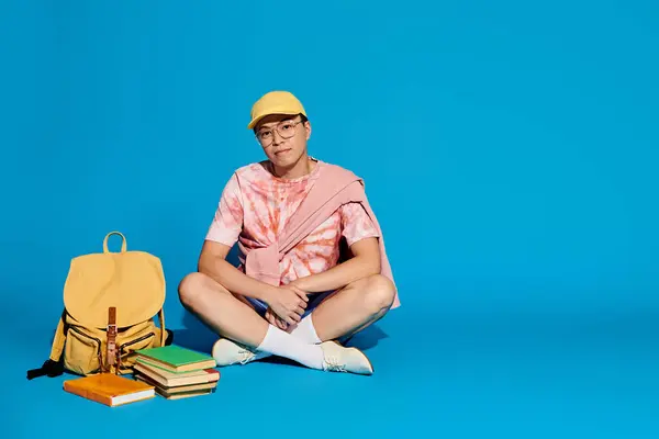 Un joven con estilo se sienta en el suelo rodeado de una mochila y libros, profundamente en el pensamiento y el estudio. - foto de stock