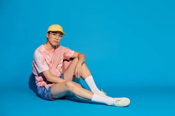 Un joven moderno y guapo con un atuendo elegante se sienta en el suelo con un sombrero amarillo contra un telón de fondo azul. - foto de stock