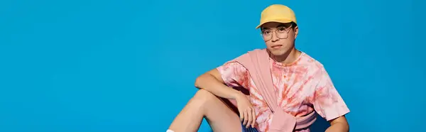 A stylish, young man sits on floor against a blue backdrop, sporting a yellow hat and trendy attire. — Stock Photo