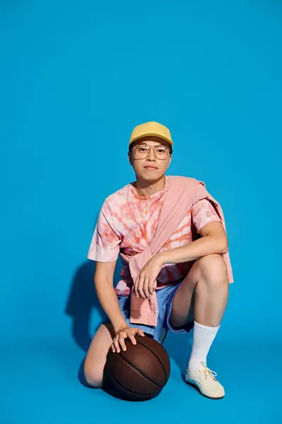 Um jovem elegante em trajes da moda sentado no chão, segurando com confiança uma bola de basquete contra um pano de fundo azul. — Fotografia de Stock
