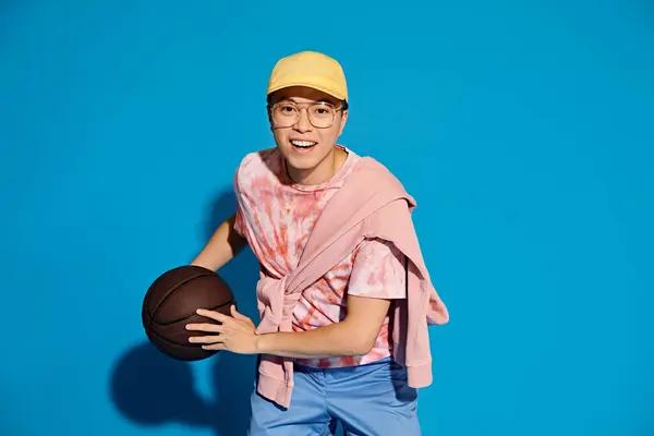 Un joven de moda sostiene enérgicamente una pelota de baloncesto en su mano derecha contra un telón de fondo azul. - foto de stock
