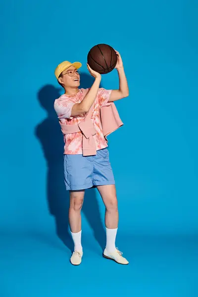 Стильный молодой человек уверенно держит баскетбольный мяч в правой руке, источая атлетизм и крутость на синем фоне.. — стоковое фото