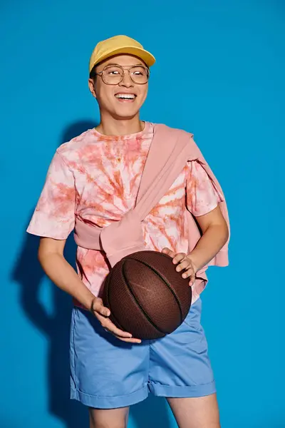 Un giovane elegante in abiti alla moda tiene energicamente un pallone da basket tra le mani su uno sfondo blu. — Foto stock