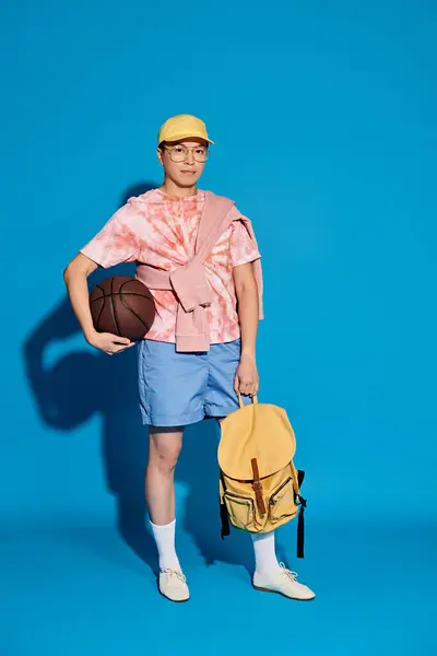 Стильный молодой человек в модном наряде держит баскетбол и рюкзак, активно позируя на синем фоне. — стоковое фото