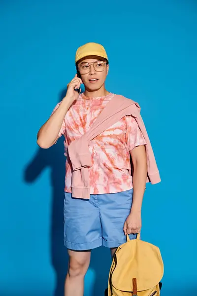 Un giovane elegante in abiti alla moda porta uno zaino e utilizza un telefono cellulare su uno sfondo blu. — Foto stock